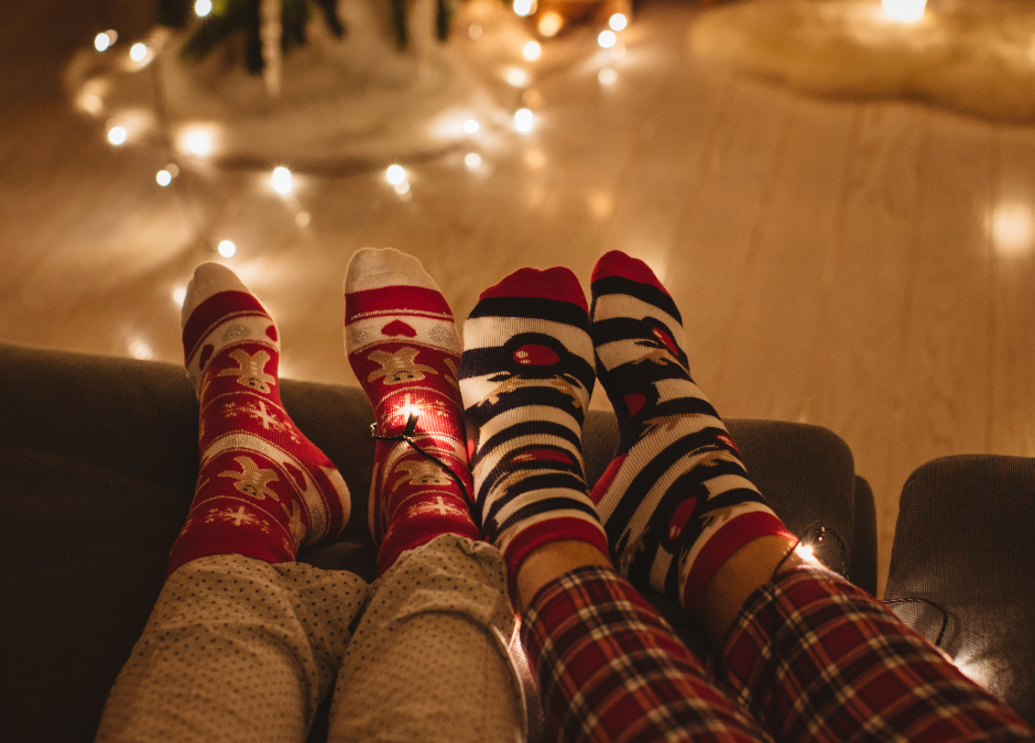 Cosy scene showing the legs from the knees down of 2 people lying on a sofa wearing pyjamas and Christmas socks. There are warm white fairy lights in the distance.